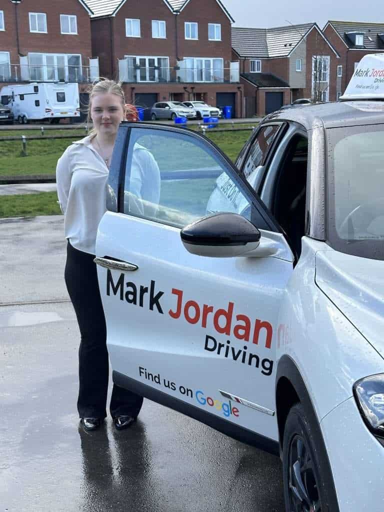 A young woman standing beside a Mark Jordan Driving School vehicle, ready for driving lessons, in a residential area with parked cars and buildings.